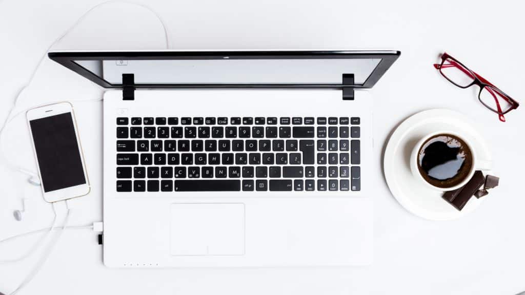 white laptop on a table next to coffee, cellphone, and reading glasses.