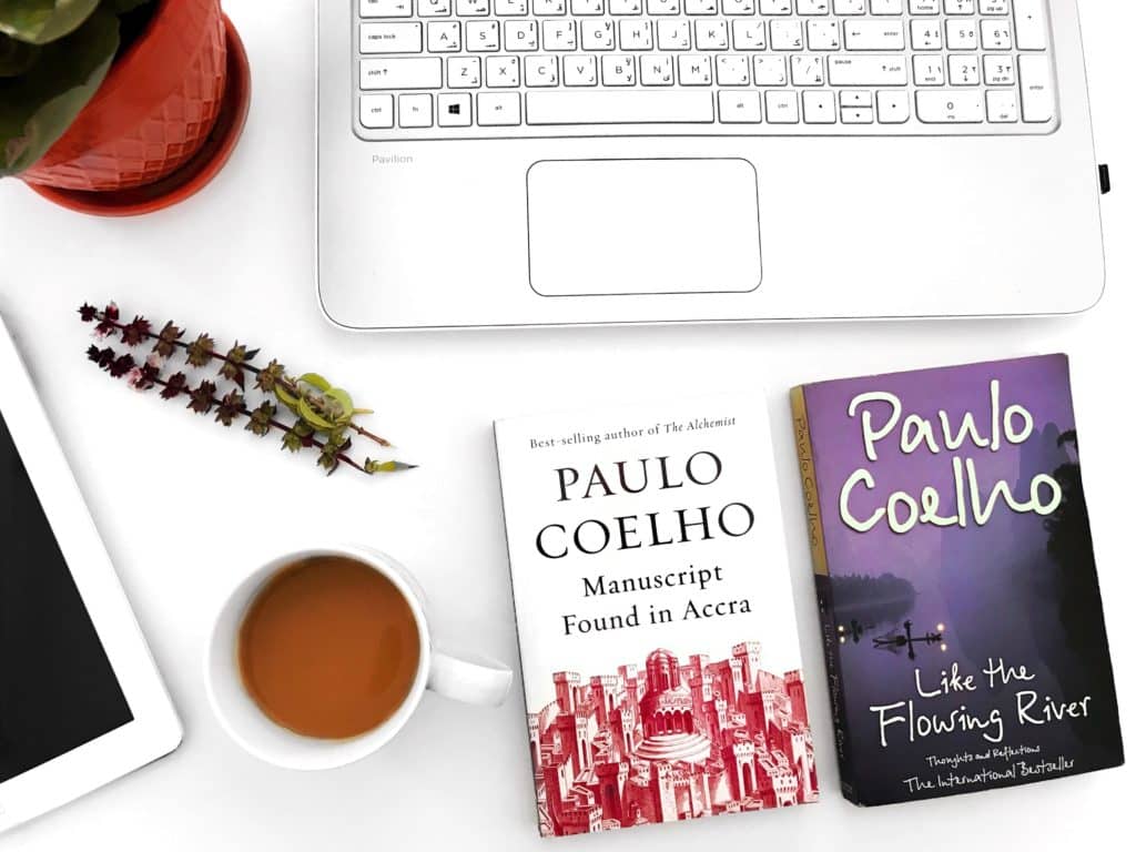 2 books next to a laptop (Like the Flowing River and Manuscript Found in Accra by Paulo Coelho), lavender, plant pot, and coffee