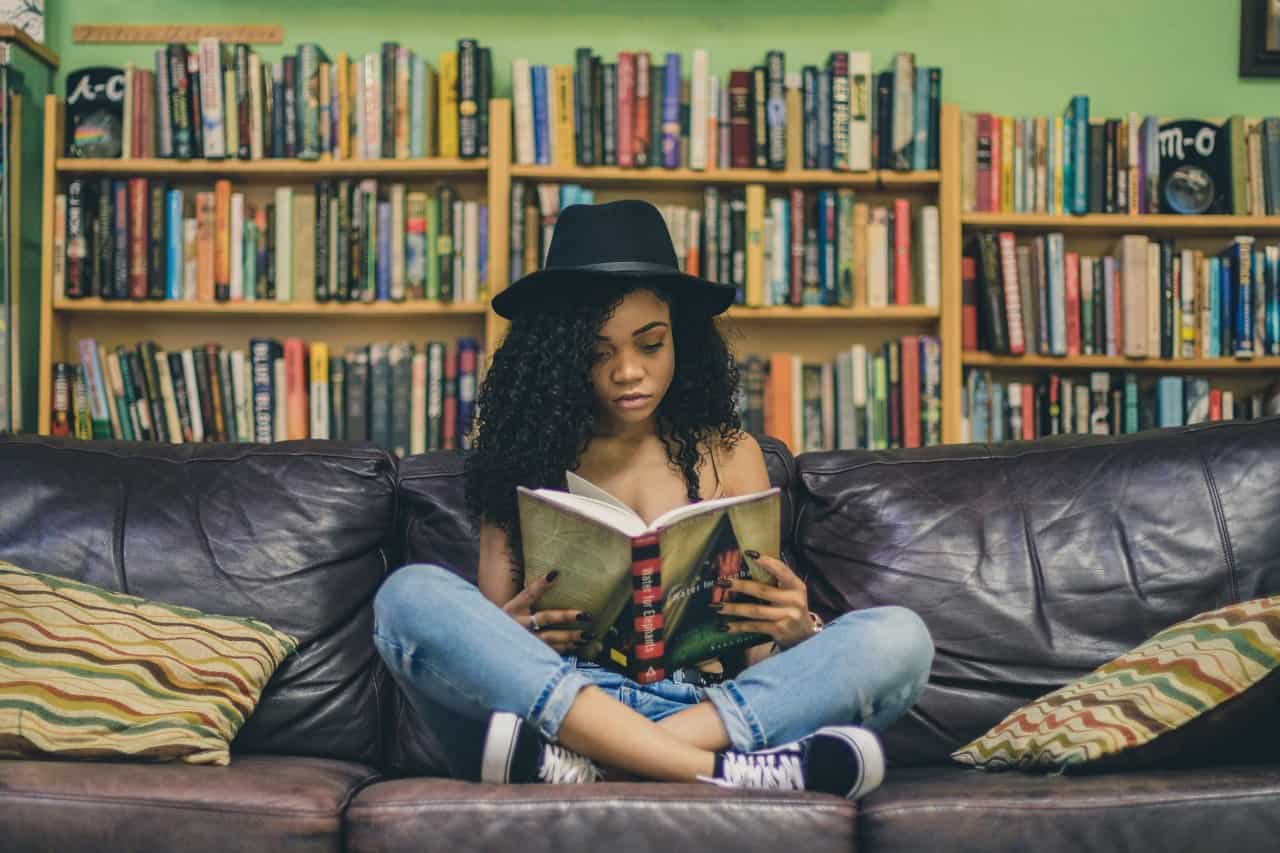 Hobbies that make money - reading - woman sitting on couch reading a book surrounded with a bookshelf in the background.