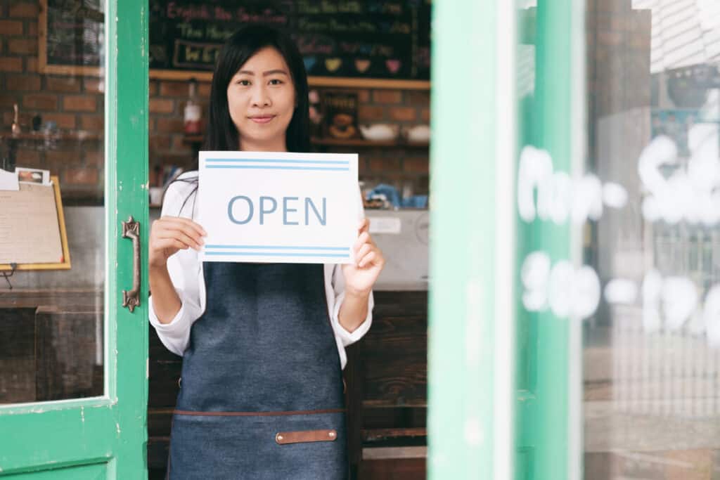support small business in post image of coffee shop and worker in smock holding "open" sign.